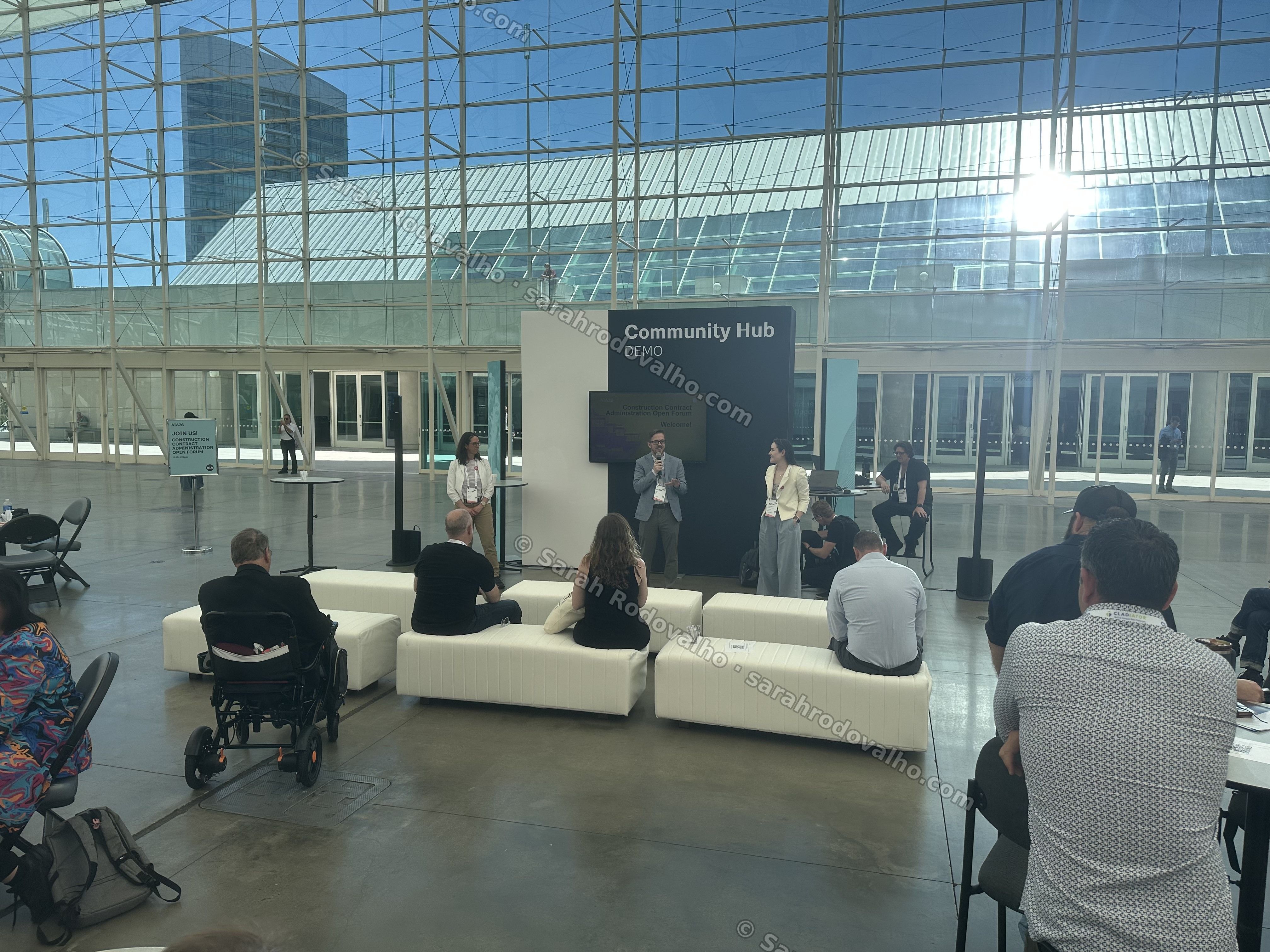Wide view of the AIA26 Community Hub 'DEMO' stage in the glass-roofed Sails Pavilion of the San Diego Convention Center, with the CCA Open Forum panel on stage and attendees seated on white benches