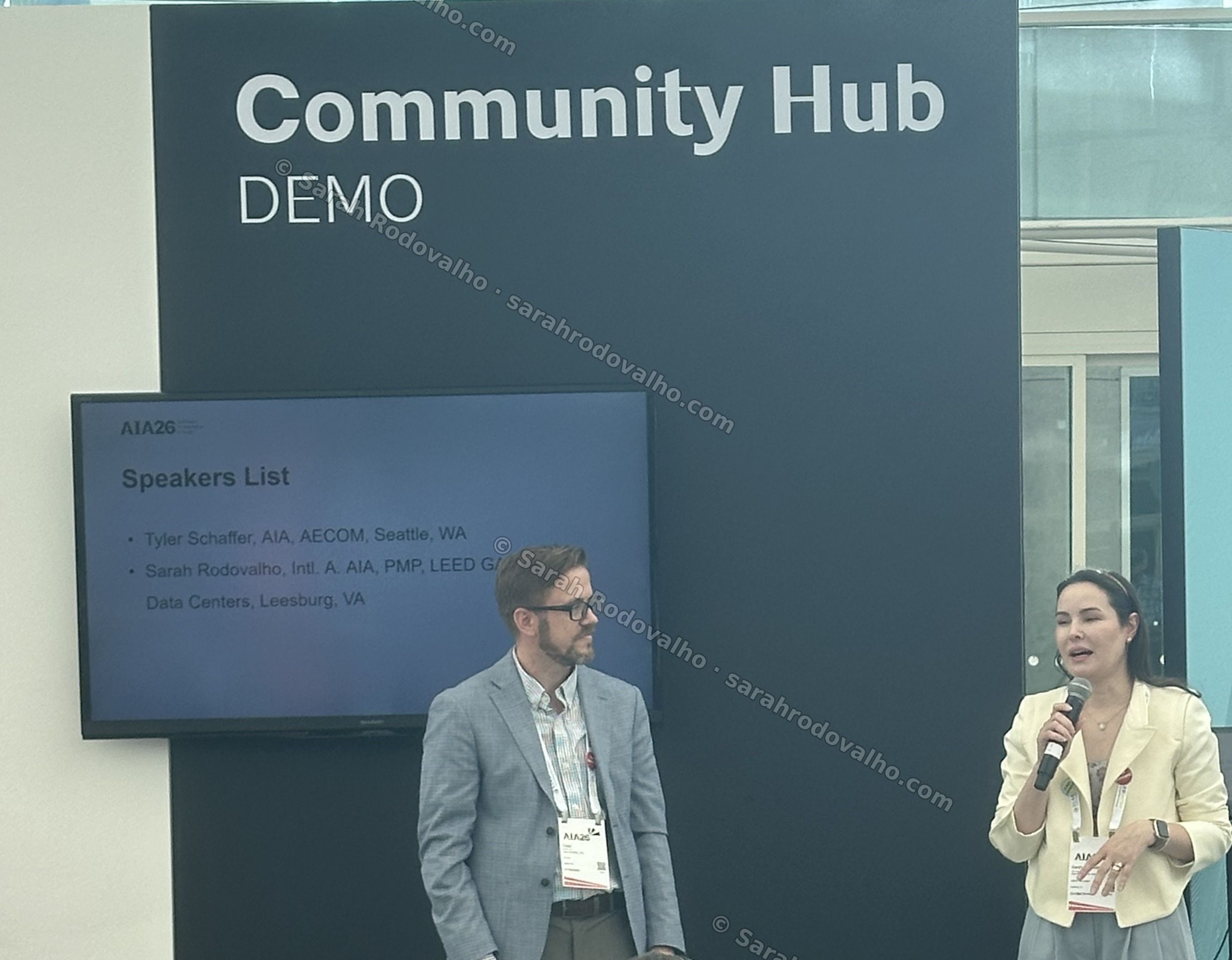 Sarah Rodovalho (right, in a cream blazer) speaking into a handheld microphone at the AIA Construction Contract Administration (CCA) Open Forum during AIA26, with co-presenter Tyler Schaffer (AIA, AECOM) at left and the on-screen 'Speakers List' slide naming both presenters — the 'Community Hub' stage at the San Diego Convention Center