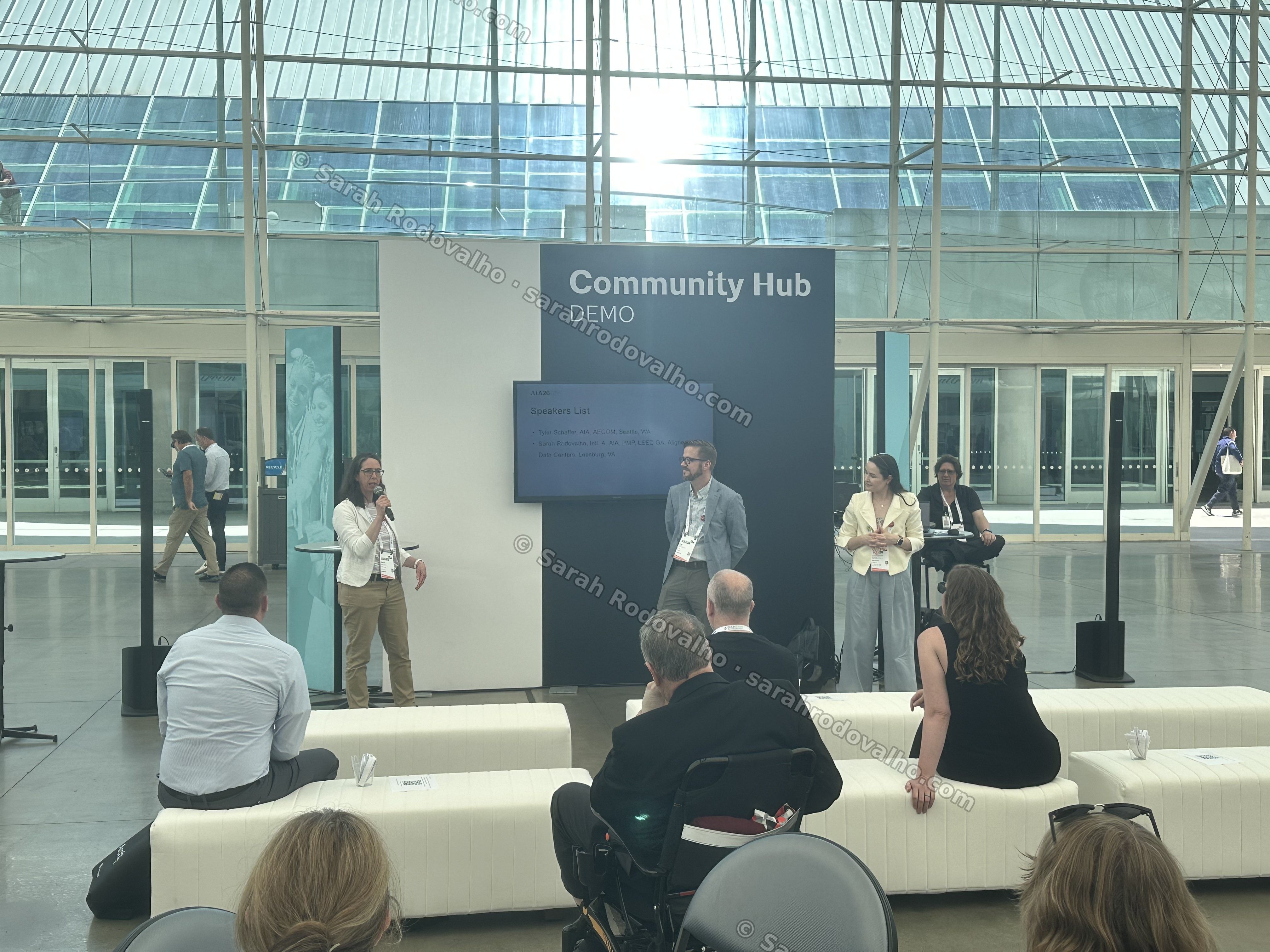 Wide view of the CCA Open Forum in session at the AIA26 Community Hub — the two presenters on stage (Sarah Rodovalho at right in a cream blazer, co-presenter Tyler Schaffer at center-left) addressing the seated audience in the glass-roofed Sails Pavilion of the San Diego Convention Center