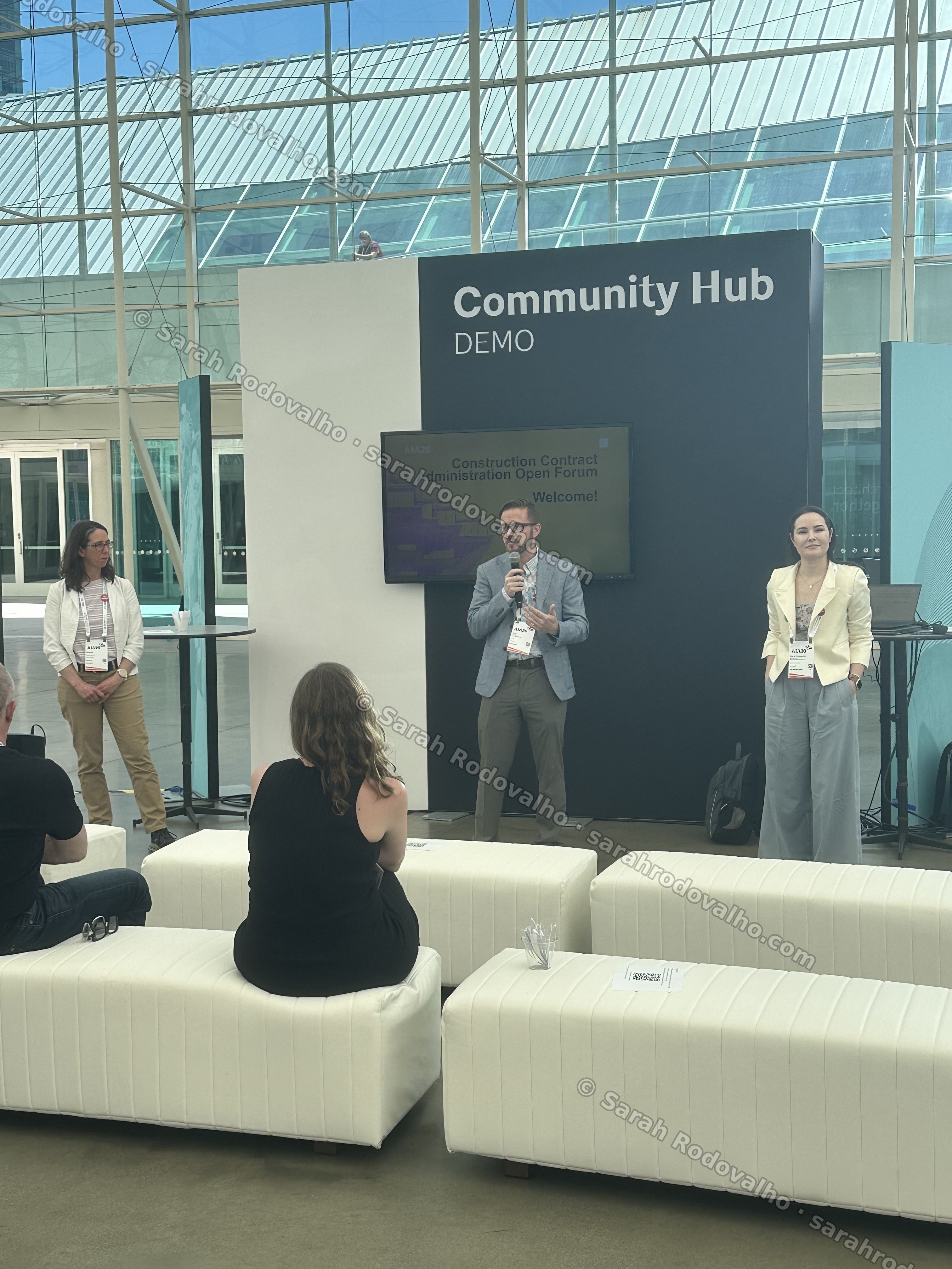 Sarah Rodovalho (right, cream blazer) on stage as the CCA Open Forum opens, a co-presenter speaking into a microphone at center, the 'Construction Contract Administration Open Forum — Welcome!' slide on screen