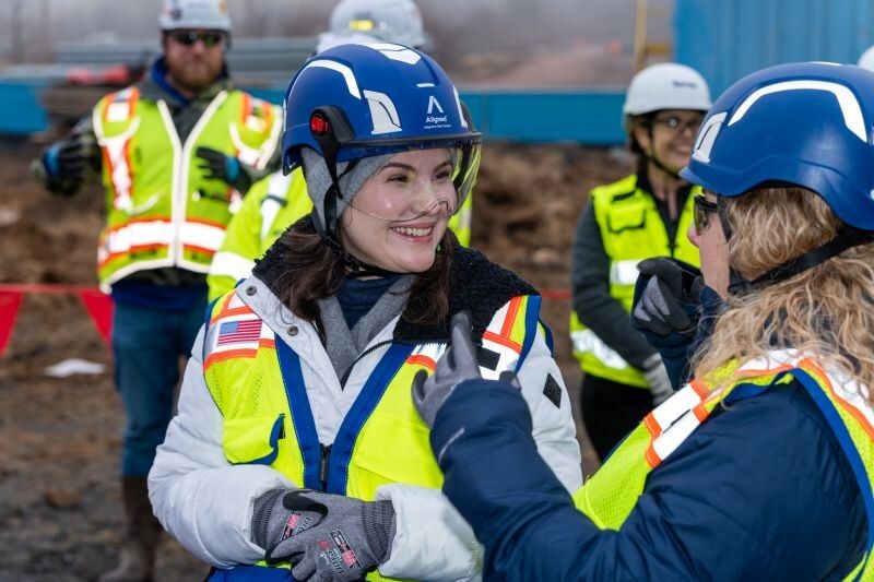 Sarah Rodovalho wearing an Aligned Data Centers branded blue hardhat and a hi-viz reflective construction vest with an American flag patch, smiling on-site at the hyperscale data-center campus during a conversation with another team member also in Aligned PPE.