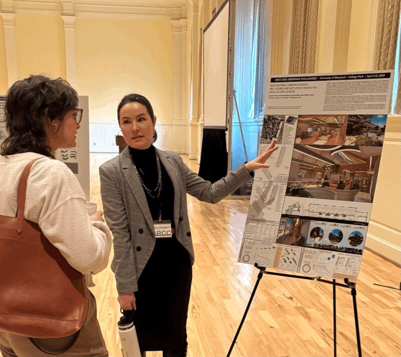 Sarah Rodovalho presenting her ARCC 2025 conference poster 'Integrating Comprehensive Wellbeing and Net-Zero Energy in Healthcare Design' at University of Maryland, College Park — gesturing toward the poster's healthcare-facility renderings and diagrams while in conversation with another attendee in the conference hall