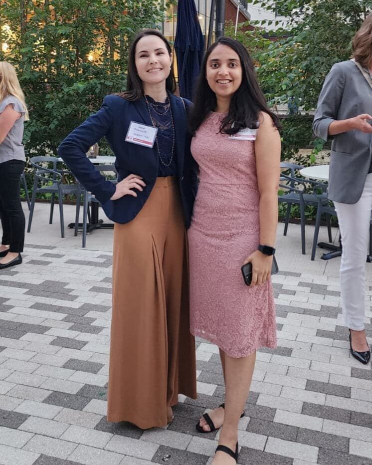 Sarah Rodovalho at a CREW Greater Philadelphia chapter event in 2022, wearing a navy blazer with a pendant necklace and her 'Sarah Rodovalho · TJU' CREW name badge, standing with another chapter member on an outdoor evening venue terrace with brick paving and café seating in the background.