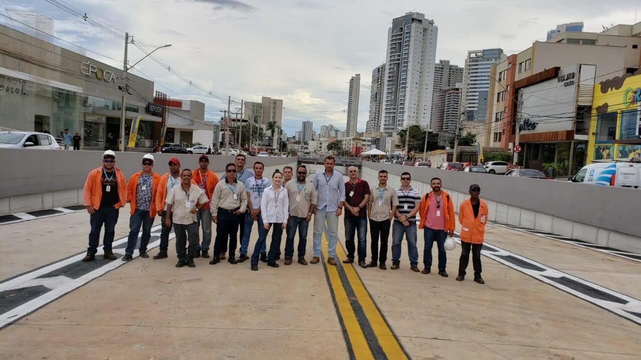 Project team on a recently paved Goiânia BRT corridor — about thirteen people, mixing construction crew in orange high-visibility vests with project-management staff wearing IDs and civilian clothing, gathered on the new busway between sloped concrete barrier walls. Goiânia's commercial skyline (ÉPOCA storefront, residential towers) is visible in the background. Sarah Rodovalho is among the project-management staff in the group.