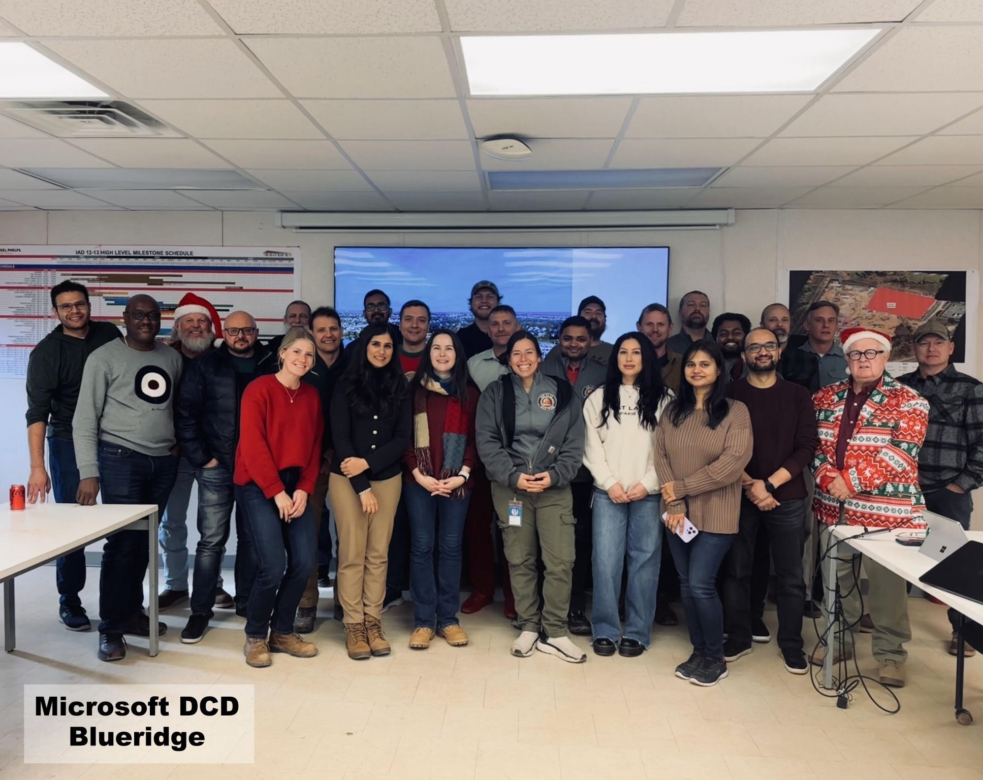 Microsoft Data Center Delivery Blueridge regional team gathered for a December 2024 group photo in a Microsoft conference room — about 25 team members including Sarah Rodovalho, with a project-milestone schedule visible on the wall behind them and a 'Microsoft DCD Blueridge' caption block overlaid on the image.