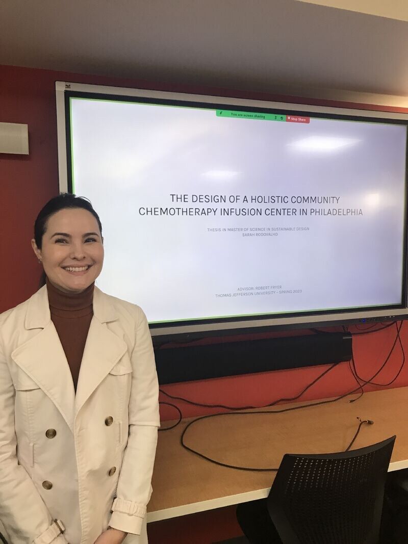 Sarah Rodovalho standing next to an interactive whiteboard displaying her M.Sc. thesis title slide: 'The Design of a Holistic Community Chemotherapy Infusion Center in Philadelphia · Thesis in Master of Science in Sustainable Design · Sarah Rodovalho · Advisor: Robert Fryer · Thomas Jefferson University · Spring 2023'.