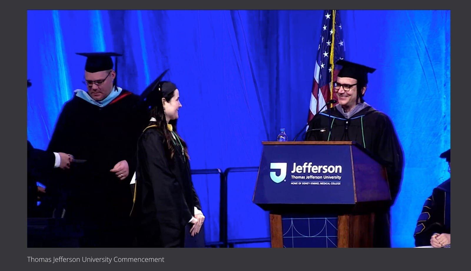 Sarah Rodovalho in cap and gown walking on stage at the Thomas Jefferson University commencement ceremony, between two academic officials in regalia; the podium displays 'Jefferson · Thomas Jefferson University · Home of Sidney Kimmel Medical College' and an American flag stands behind the stage.