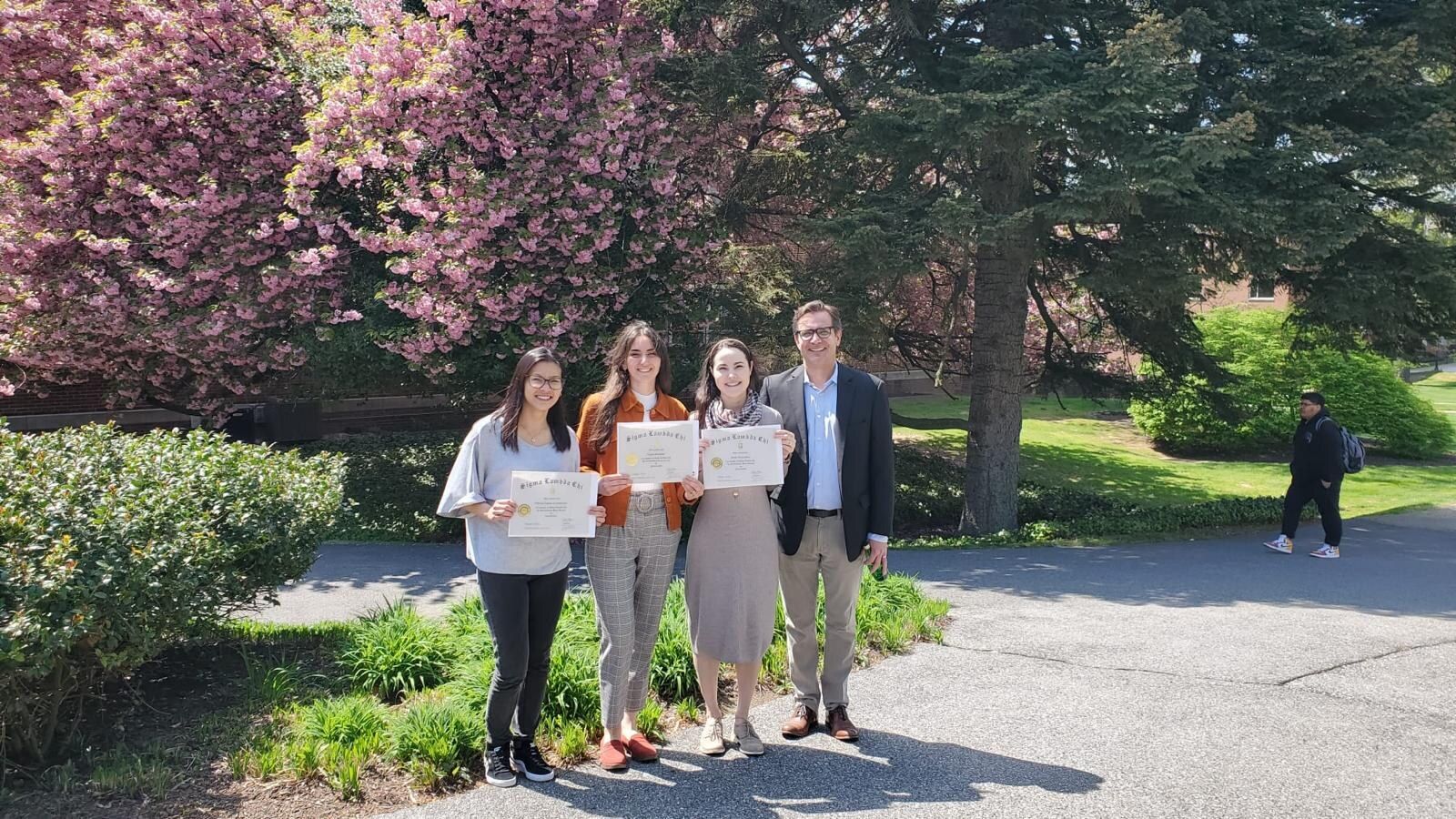 Sigma Lambda Chi induction certificate ceremony — four people outdoors among cherry blossoms on Thomas Jefferson University campus: three newly-inducted student members each holding a Sigma Lambda Chi certificate, and the sponsoring faculty member on the right. Sarah Rodovalho stands third from the left in a long tan dress.