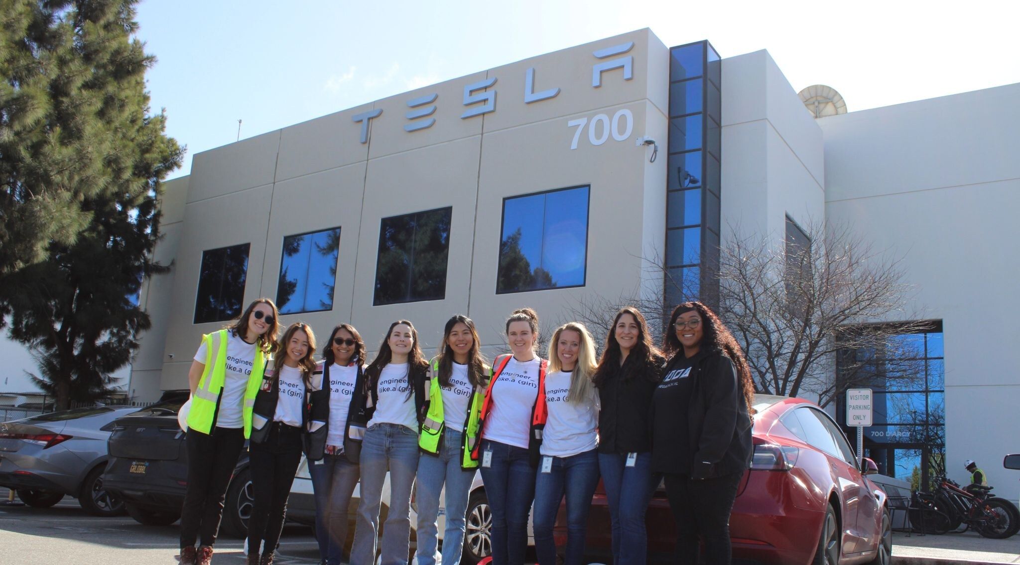 Eight women from Tesla in matching 'engineer like a girl' t-shirts and hi-viz vests, standing in front of the Tesla 700 building entrance — facilitators of the 2024 Introduce a Girl to Engineering Day at the Fremont Megafactory.