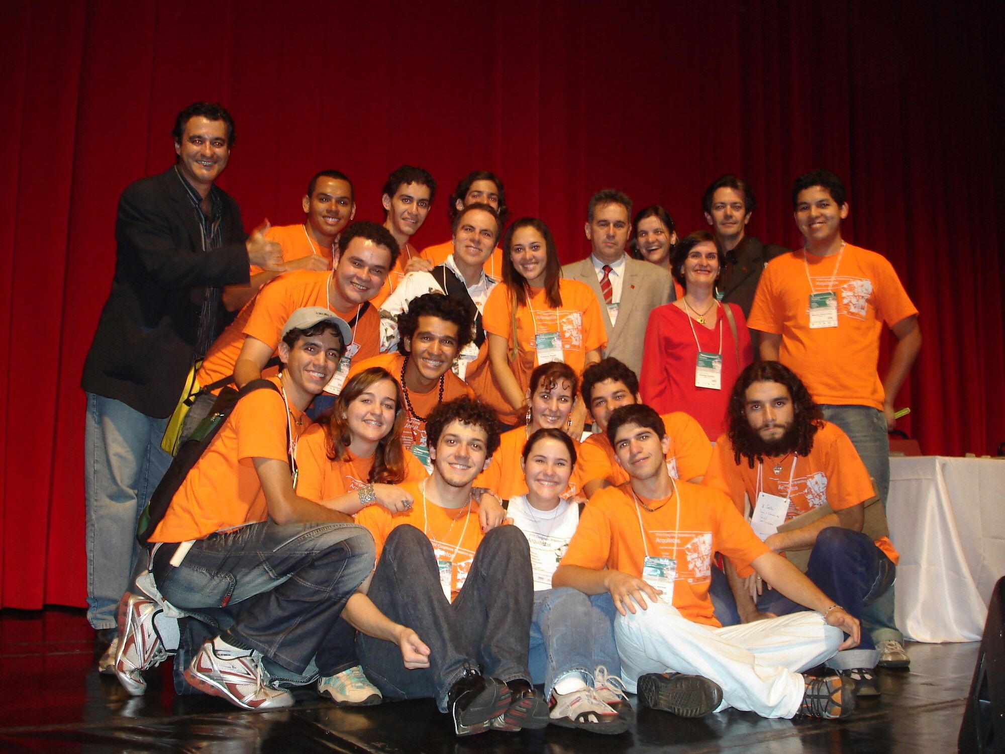 Stage group portrait of about seventeen members of the XVIII Brazilian Architects Congress organizing support team, taken on stage with a red curtain backdrop on 2006-10-14. The team wears matching orange organizing t-shirts; Sarah Rodovalho, then an undergraduate architecture student at UEG, kneels at the center front in a white t-shirt.