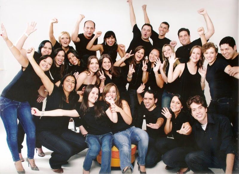 Studio class portrait of Sarah Rodovalho's UEG Architecture and Urban Planning graduating cohort — about twenty graduates coordinated in black tops and jeans against a white seamless backdrop, posed with arms raised, peace signs, and casual smiles in the Brazilian 'foto da turma' graduating-class tradition.